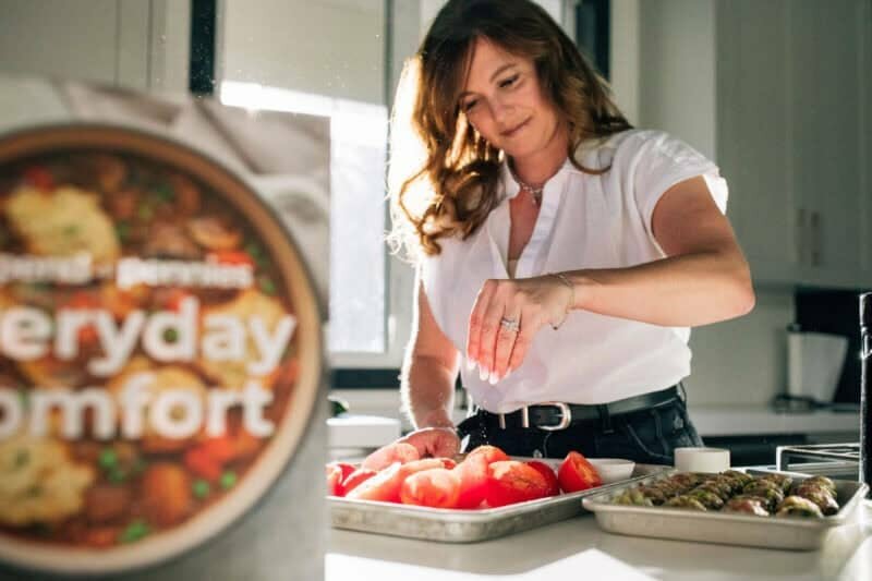 Holly Nilsson seasoning tomatoes with her cookbook Everyday Comfort in the foreground